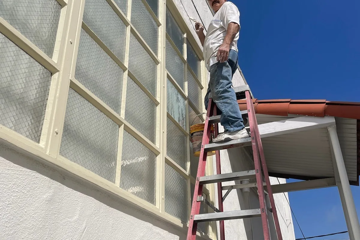 Tony's Painting crew member on a ladder painting a white stucco wall beside a large window, San Diego County